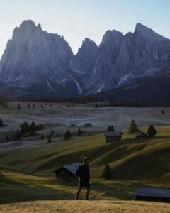 A person gazing at a stunning mountain range from a grassy hill at sunrise.