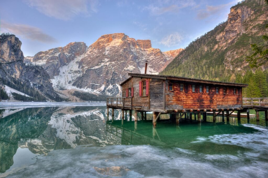 A serene lakeside cabin reflecting on water surrounded by majestic mountains.