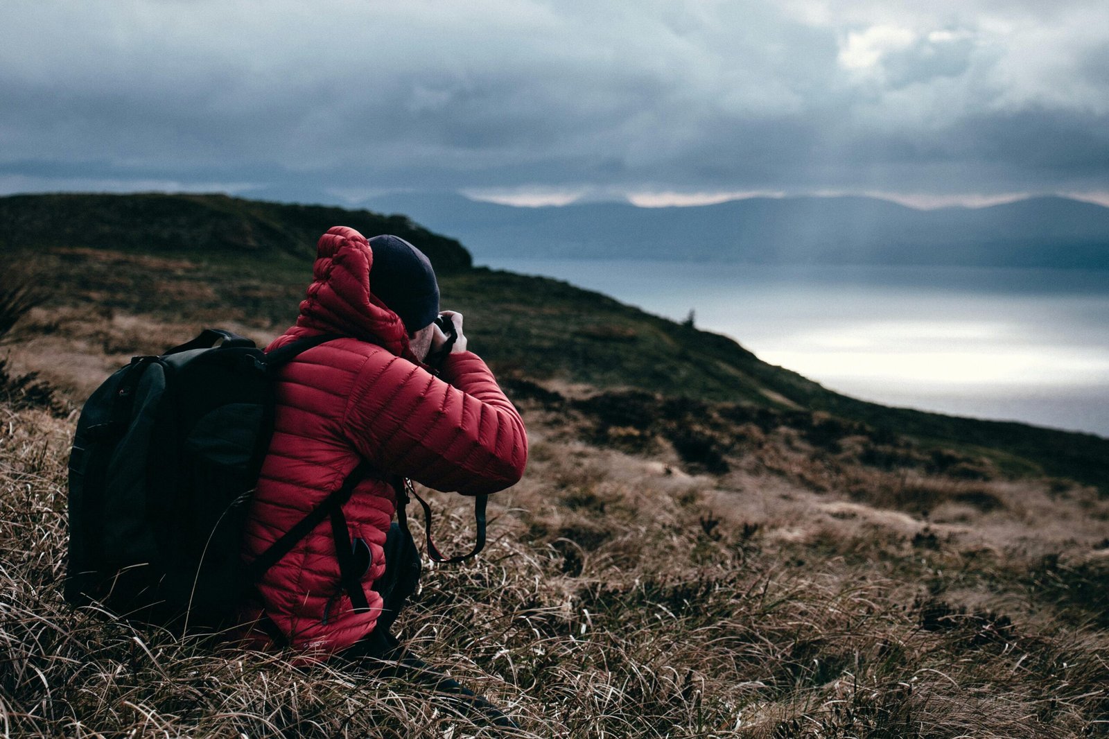 A hiker in a red jacket photographs a scenic mountain sunset by the sea.