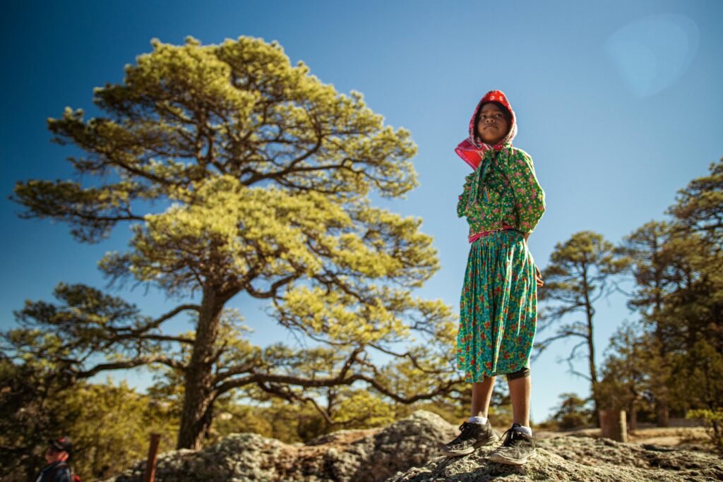 Raramuri girl standing on a rock in traditional clothing with a forest background under clear skies.