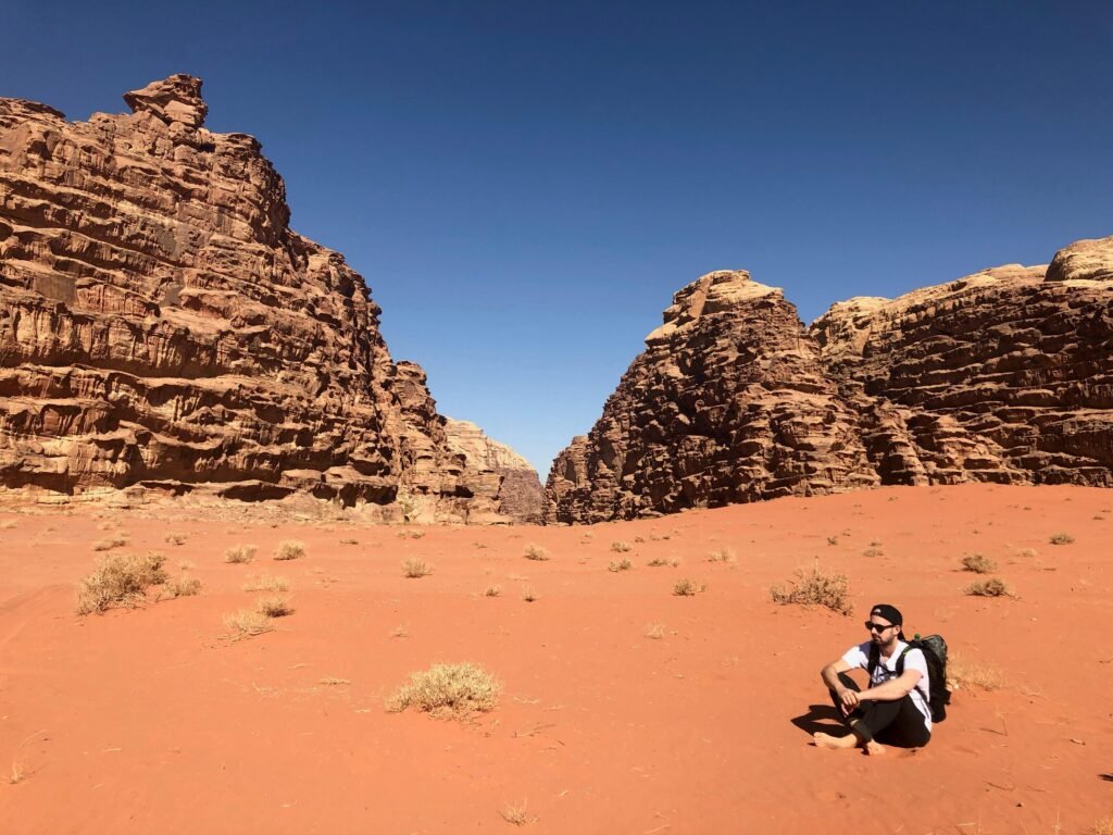 A hiker with a backpack explores the arid desert canyons of Wadi Rum under a clear blue sky.