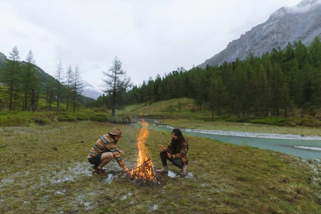 A couple enjoys camping by the riverside with a bonfire in scenic mountain terrain.